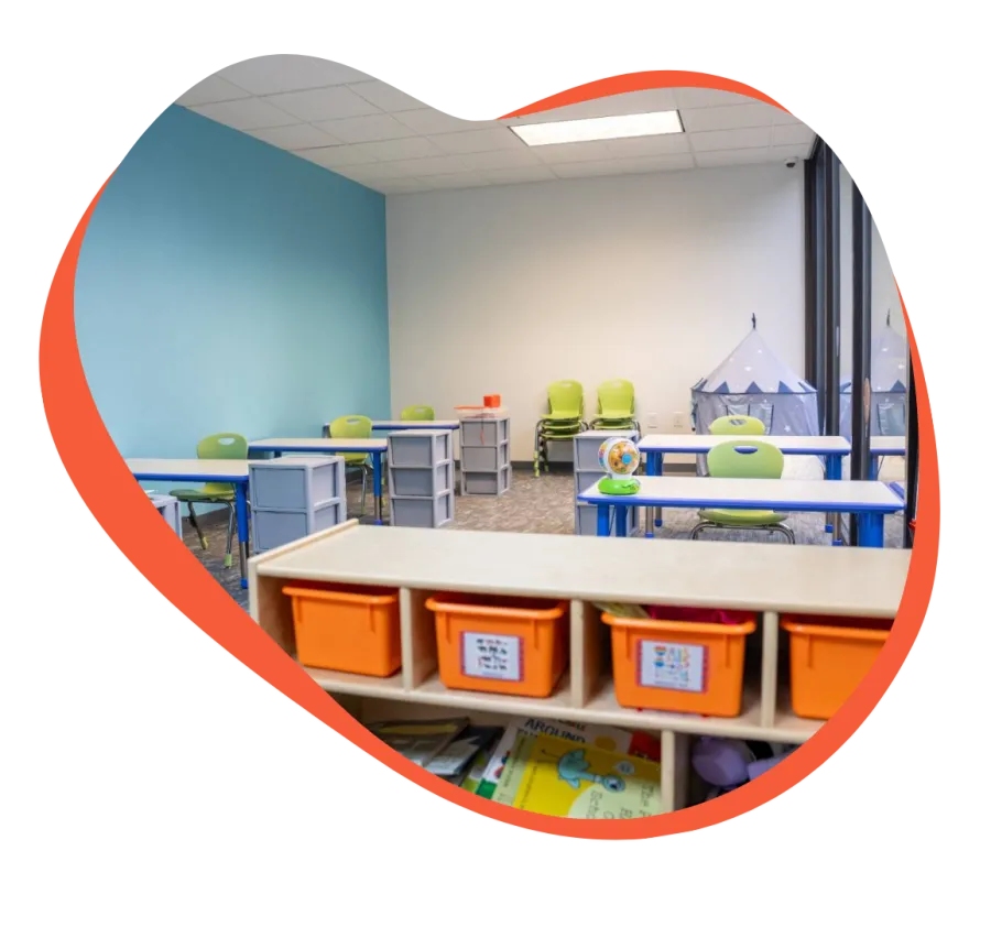 Bright classroom with blue and white walls, green chairs, orange storage bins, and children's play tent.