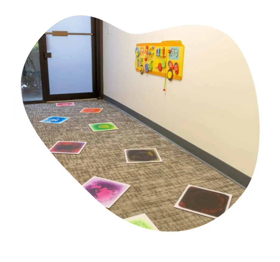 Colorful sensory play mats arranged on carpeted floor leading to a play activity board on the wall in a bright hallway.