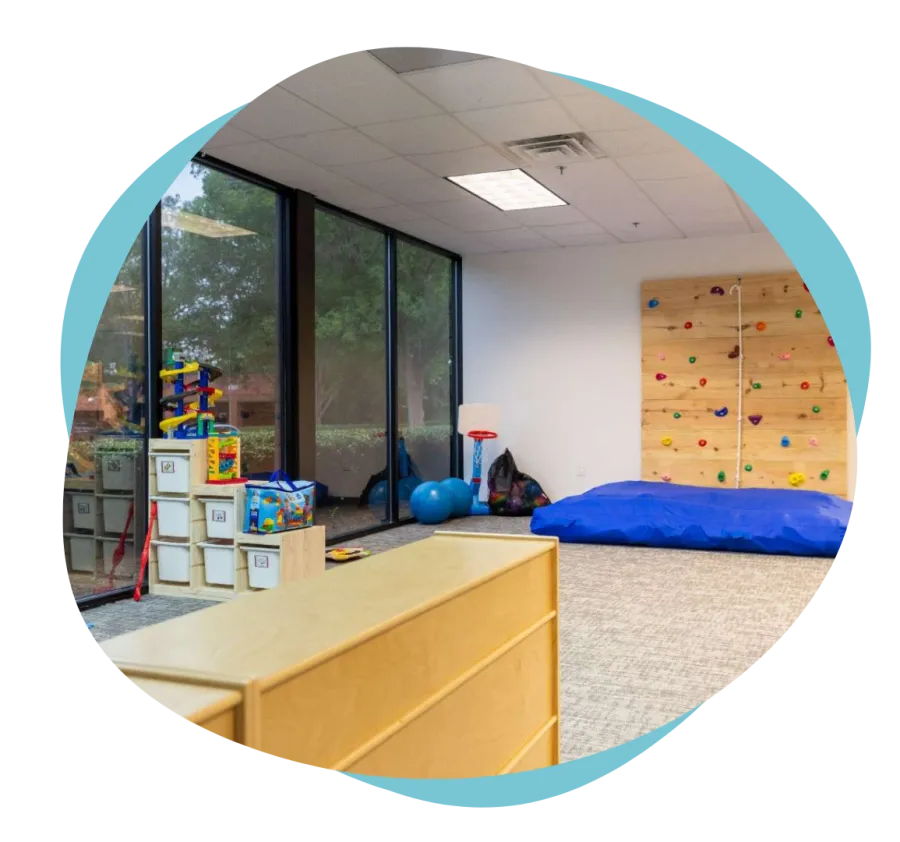 Bright playroom with climbing wall, soft mat, basketball hoop, toys, and large windows overlooking greenery.
