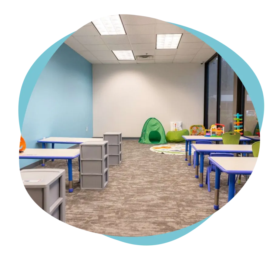 Therapist sitting with clipboard interacting with young boy holding a toy ball in a colorful playroom.