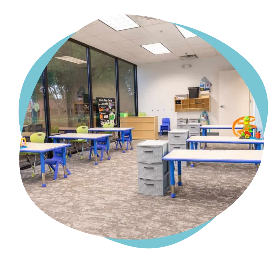 Bright classroom with blue and green chairs, tables, storage drawers, and large windows overlooking trees.
