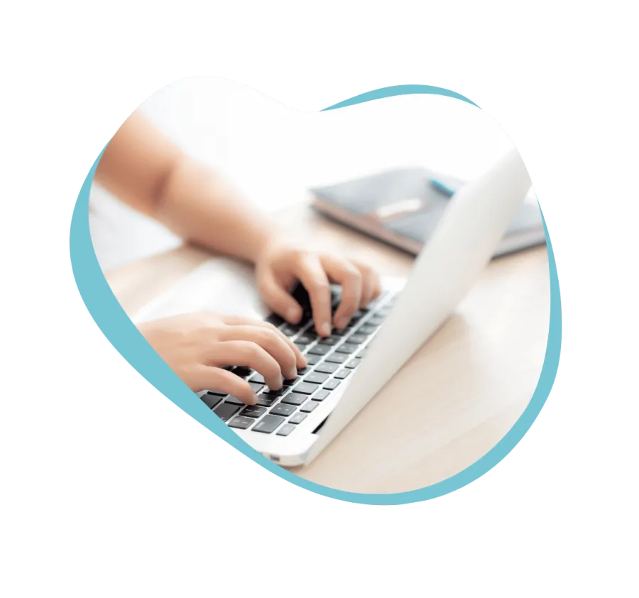 Person typing on a laptop keyboard at a wooden desk with a notebook and pen in the background