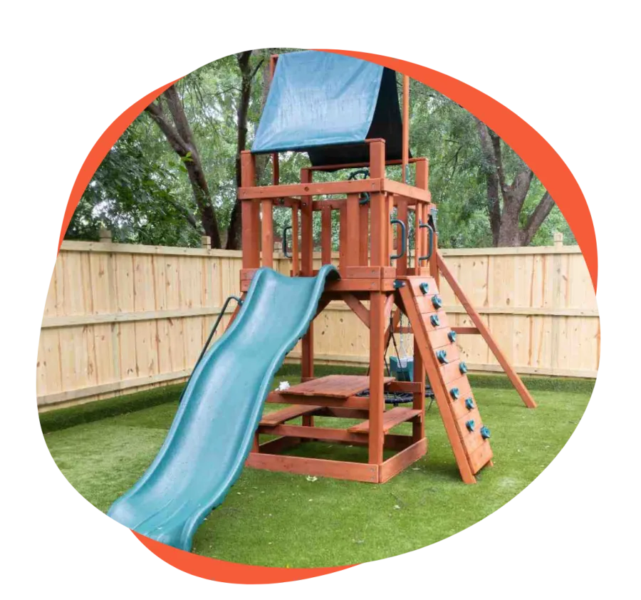 Wooden outdoor playground set with blue slide, climbing wall, and canopy on green grass in fenced backyard.
