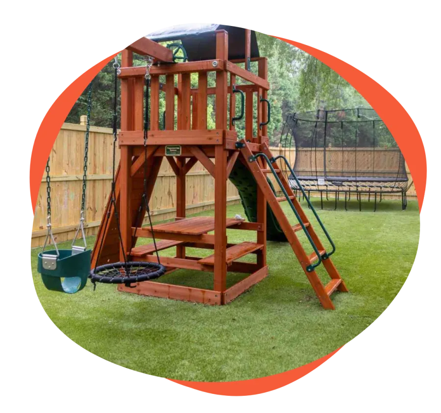 Wooden playground set with swings, slide, picnic table, and trampoline on green lawn in fenced backyard.