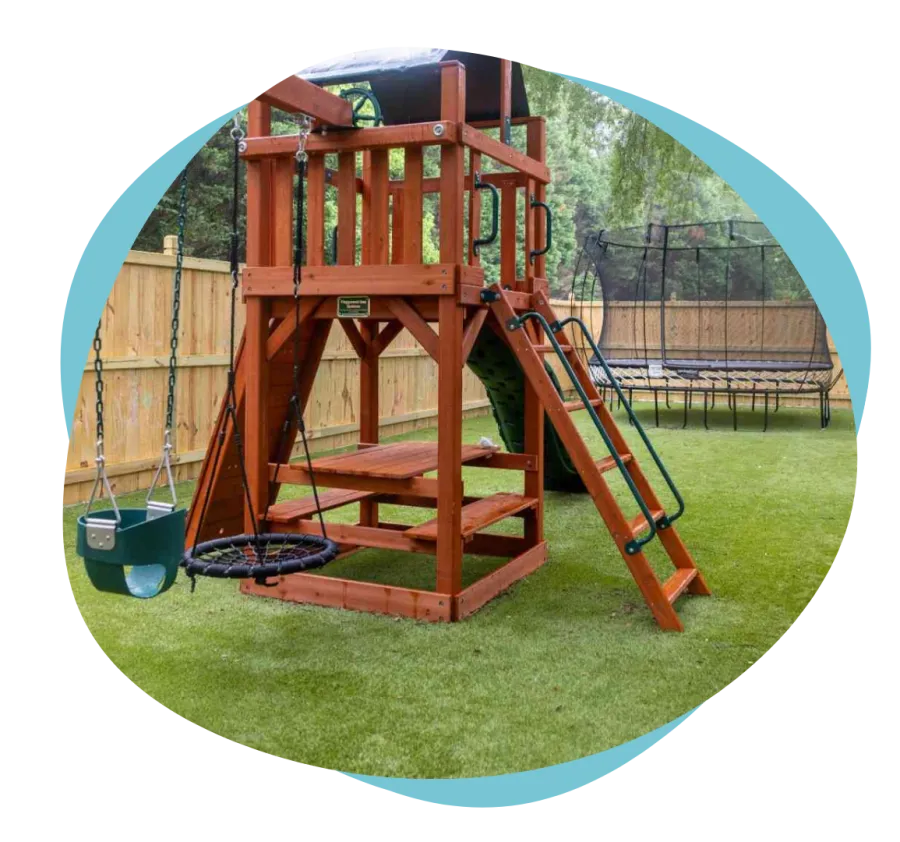 Wooden backyard playset with swings, climbing ladder, slide, picnic table, and trampoline on green grass.