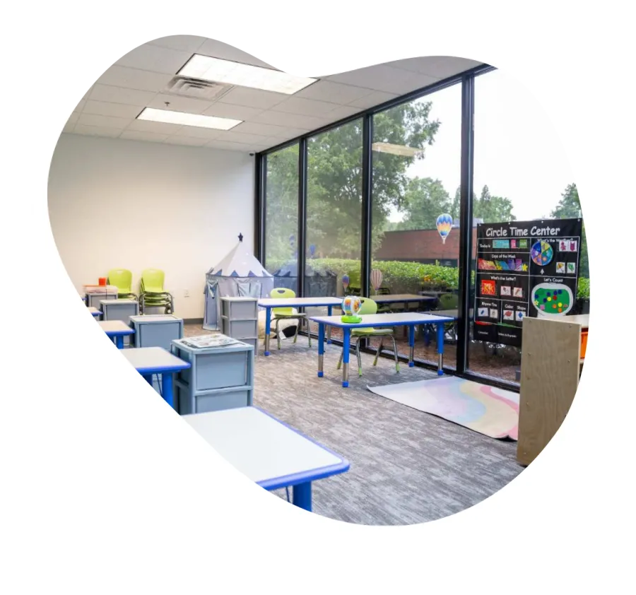 Bright early childhood classroom with colorful chairs, learning posters, play tent, and large windows overlooking greenery.