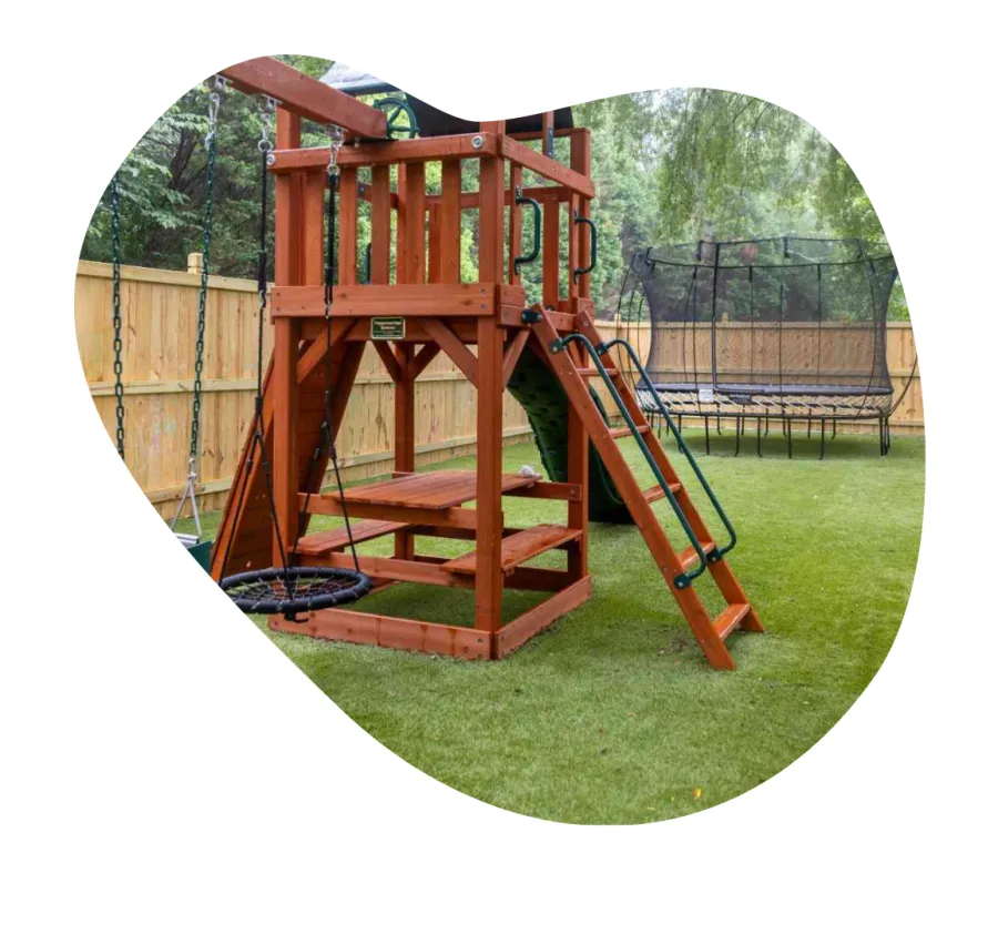 Wooden backyard playset with swing, slide, climbing wall, and picnic bench on green lawn near wooden fence.