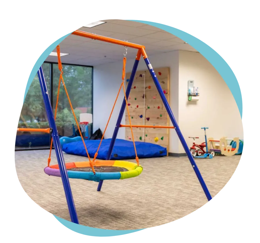 Indoor children's play area with a colorful round swing, climbing wall, and various ride-on toys.