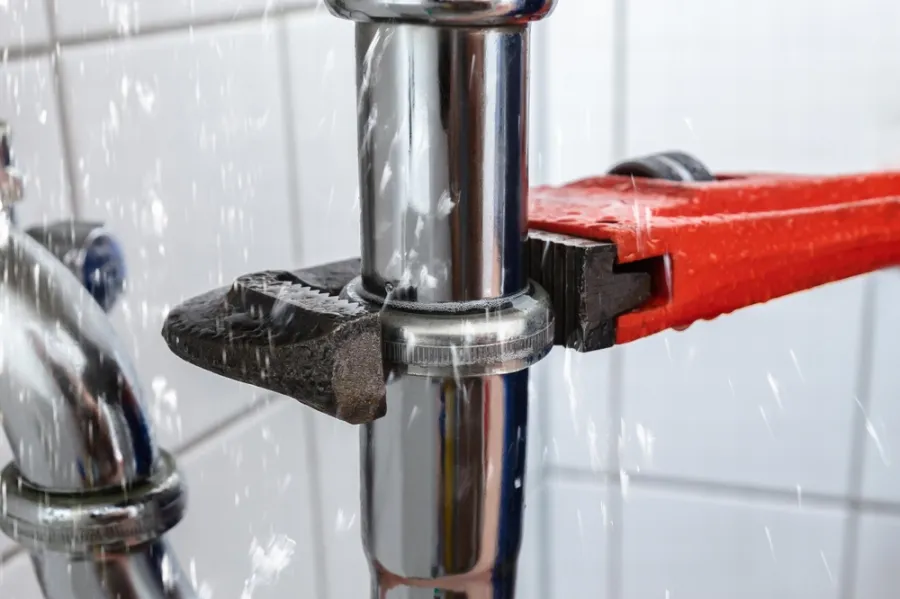 Close-up of a plumber's wrench tightening a metal pipe with water spraying from a leak.
