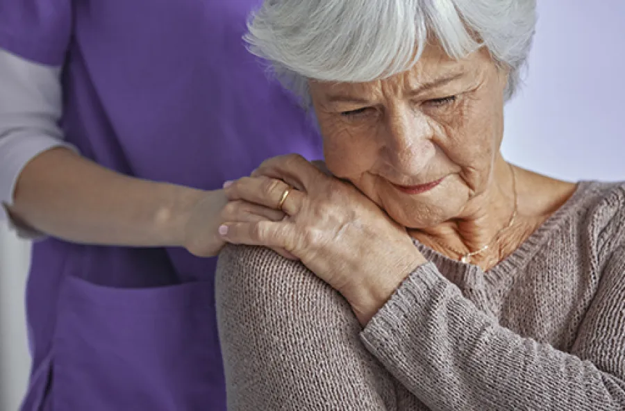 Senior woman receiving supportive shoulder touch from caregiver wearing purple uniform indoors