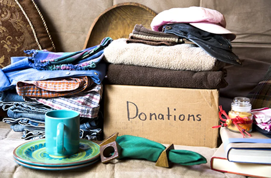 Box labeled donations with folded clothes, hats, books, a mug, and a plate arranged for charity giving.