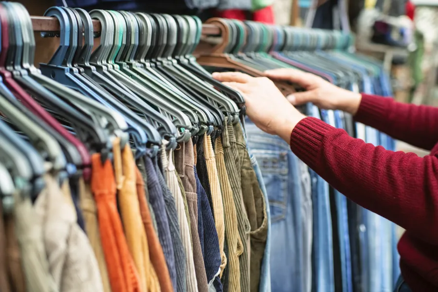 Person browsing a rack of various colored pants and jeans hanging on metal hangers in a clothing store.