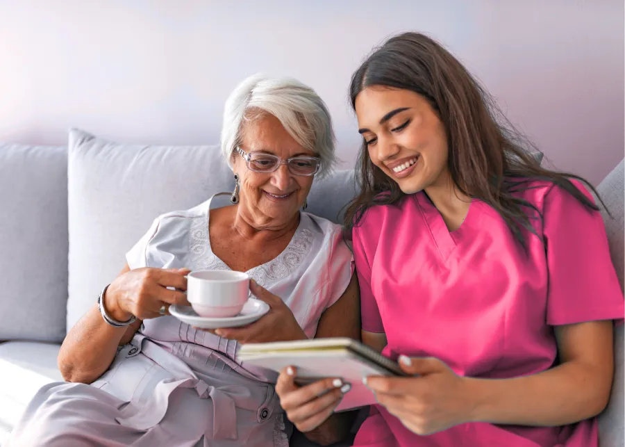 Elderly woman and young nurse smiling together while reading a book on a couch, elderly woman holding a teacup.