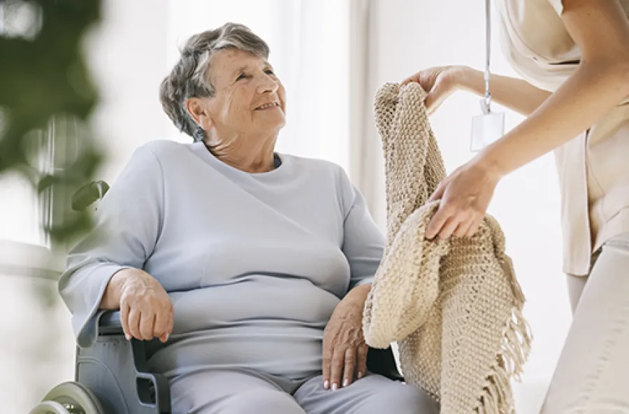 Elderly woman in wheelchair smiling at caregiver offering a knitted blanket in a bright room.