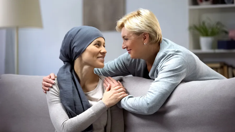 Woman wearing headscarf comforted by friend on couch in cozy living room setting.