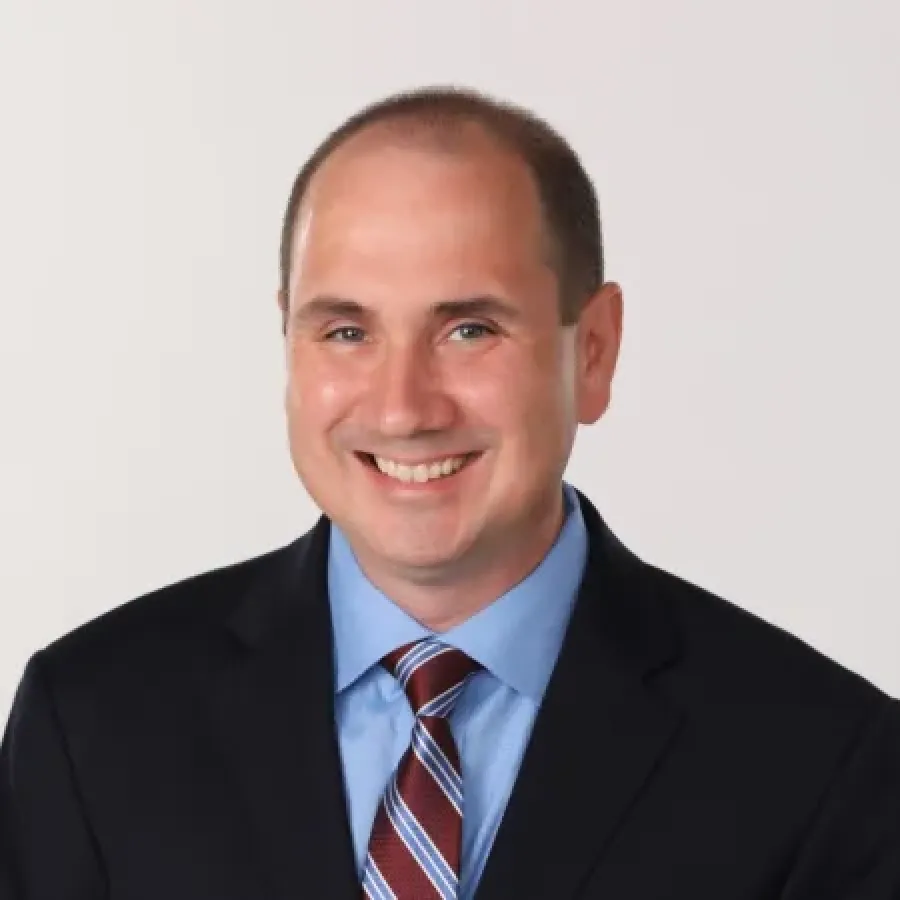 Smiling man in black suit, blue shirt, and striped tie against a plain light background