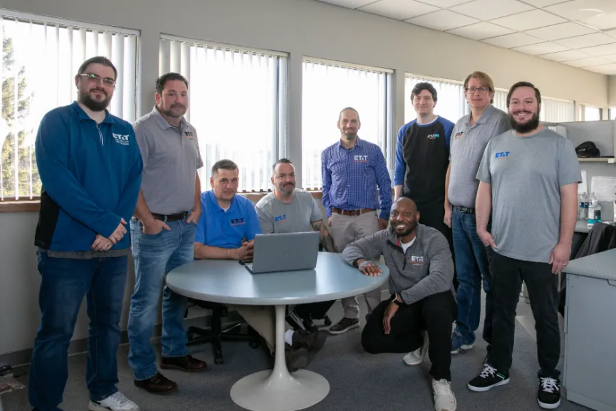 Group of professionals in casual attire gathered around a table with a laptop in an office setting.