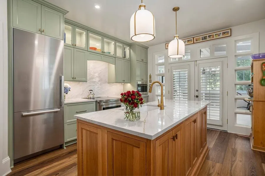 Modern kitchen with green cabinets, wooden island, marble countertop, stainless steel fridge, and pendant lighting.