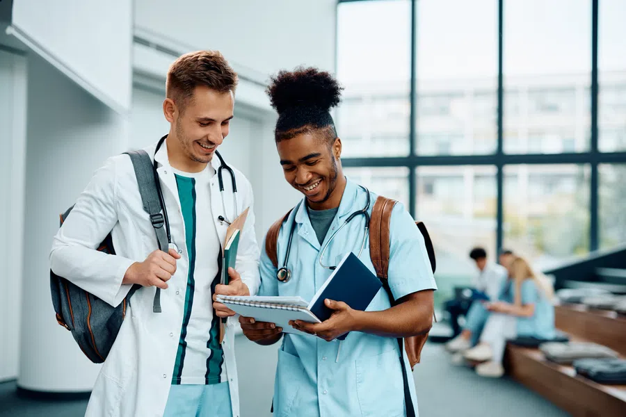 Two medical students in scrubs and lab coat smiling while reviewing notes together in a bright campus lobby.