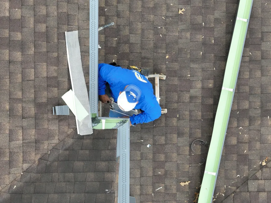 A worker in blue shirt and white cap installs green gutters on a brown shingled roof from above.