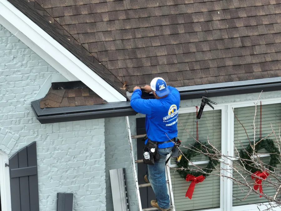 Worker in blue uniform installs or repairs gutter on a residential house with holiday wreaths on windows.