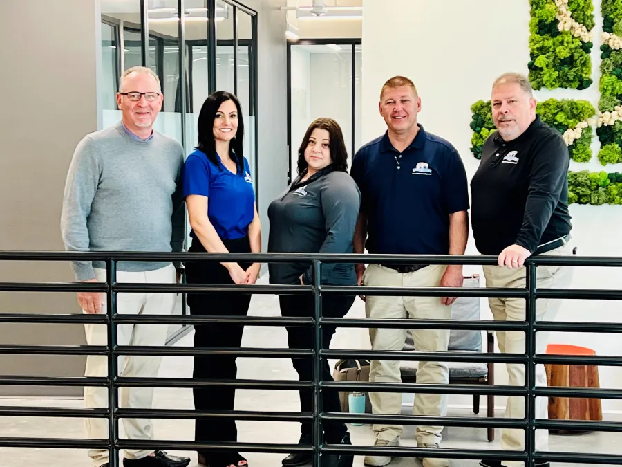 Five professionals standing indoors behind a black railing in a modern office setting with green wall art.