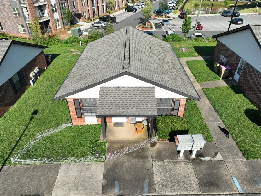 Aerial view of a single-story brick house with gray roof, small fenced yard, and nearby parking lot at apartment complex.