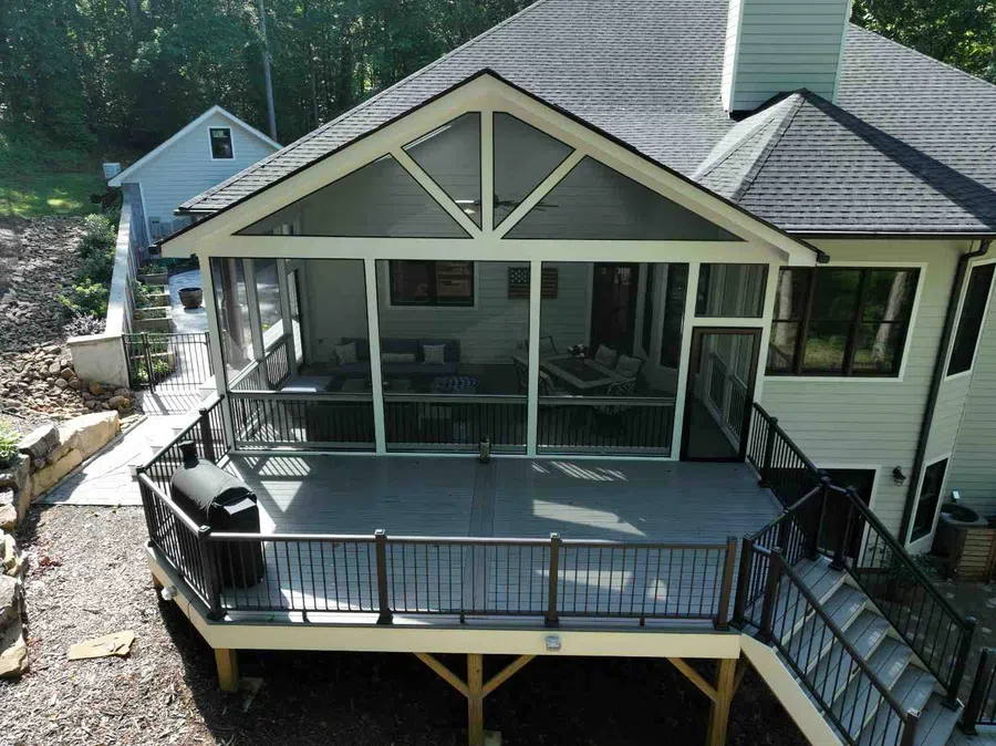 Elevated deck with screened porch attached to a white house overlooking a wooded backyard area.