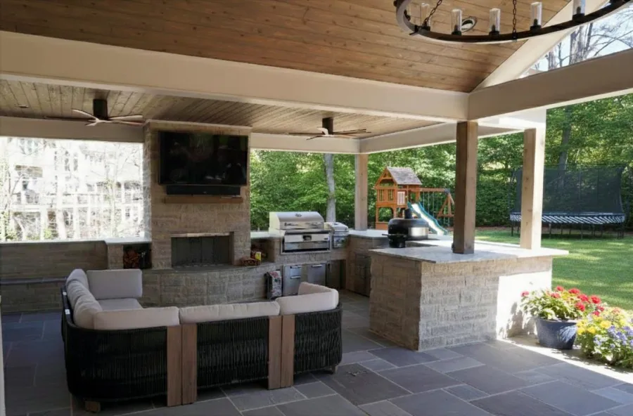 Covered outdoor patio with fireplace, TV, grill, cushioned seating, playset, and garden under wooden ceiling.