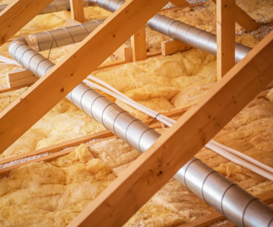 Yellow fiberglass insulation installed between wooden attic beams with silver ventilation ducts.