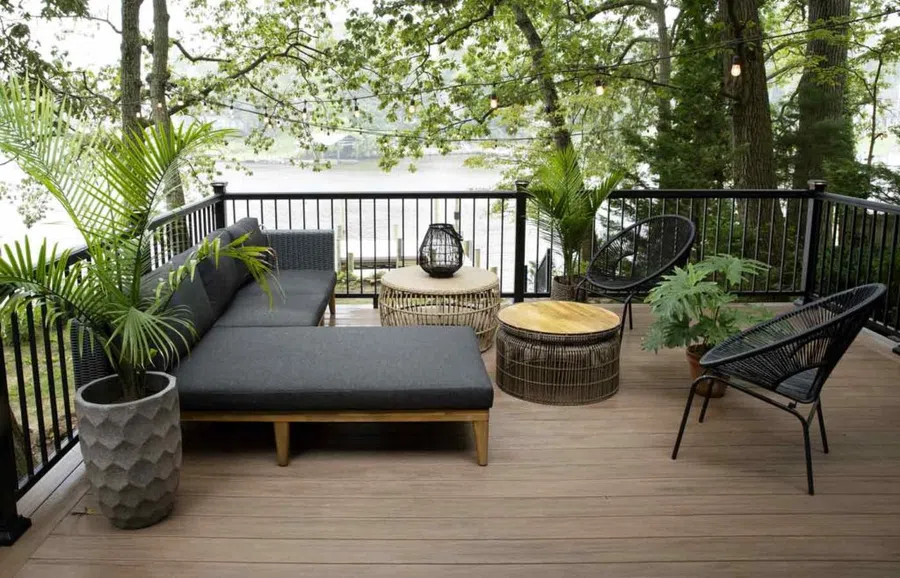 Modern outdoor seating area on wooden deck with gray sofa, round tables, black chairs, potted plants, and river view