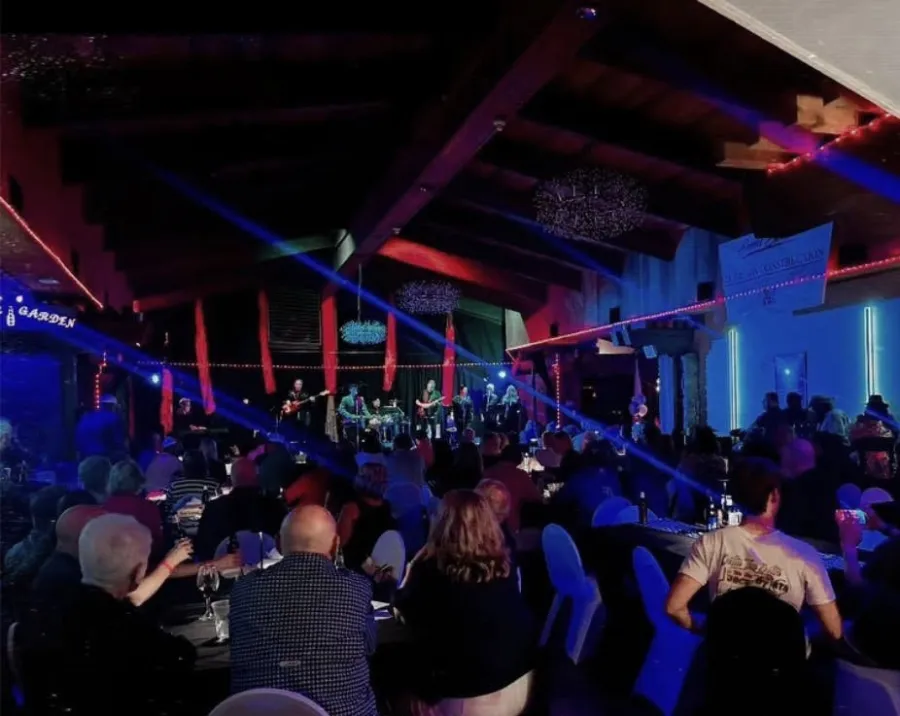 Crowded indoor concert venue with a band performing on stage under colorful red and blue lights.