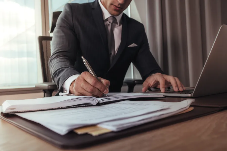 Businessman in a suit writing on documents at a desk with a laptop and paperwork in an office setting.