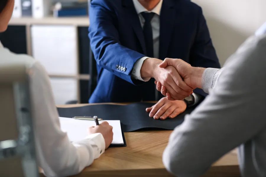 Business professionals shaking hands during a meeting with a clipboard and pen on the table.