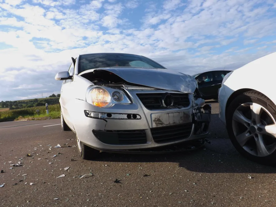 Silver car with extensive front-end damage after a collision, debris scattered on the road under a partly cloudy sky