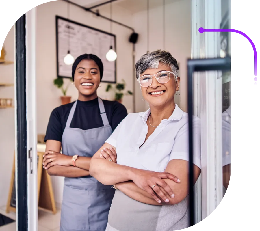 Two smiling cafe employees wearing aprons standing confidently with arms crossed inside their coffee shop.