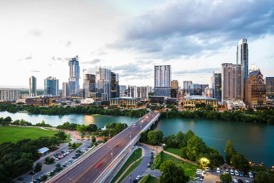 Aerial view of a city skyline with tall buildings, a river, and a bridge under a partly cloudy sky at dusk.