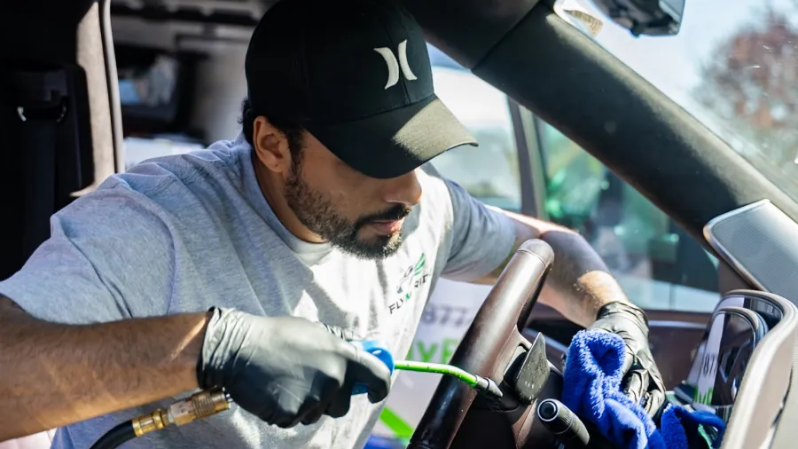 Man wearing gloves cleaning and detailing the interior steering wheel of a car with tools and cloth.