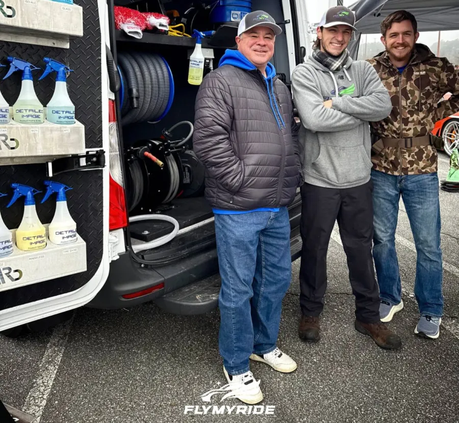 Three men standing next to a van with car detailing supplies and equipment in an outdoor setting.