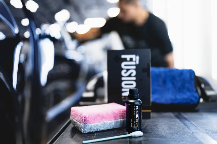 Bottle of Fusion Plus ceramic coating with applicator towels on a table beside a shiny black car and blurred technician.