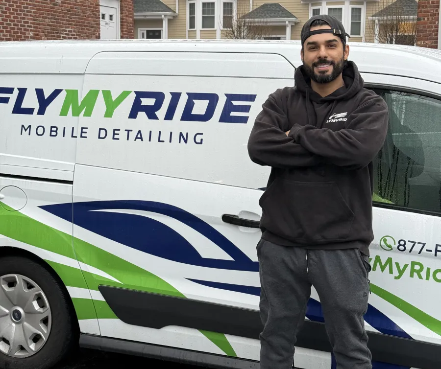 Smiling man in black hoodie standing with arms crossed by Fly My Ride mobile detailing van in residential area.
