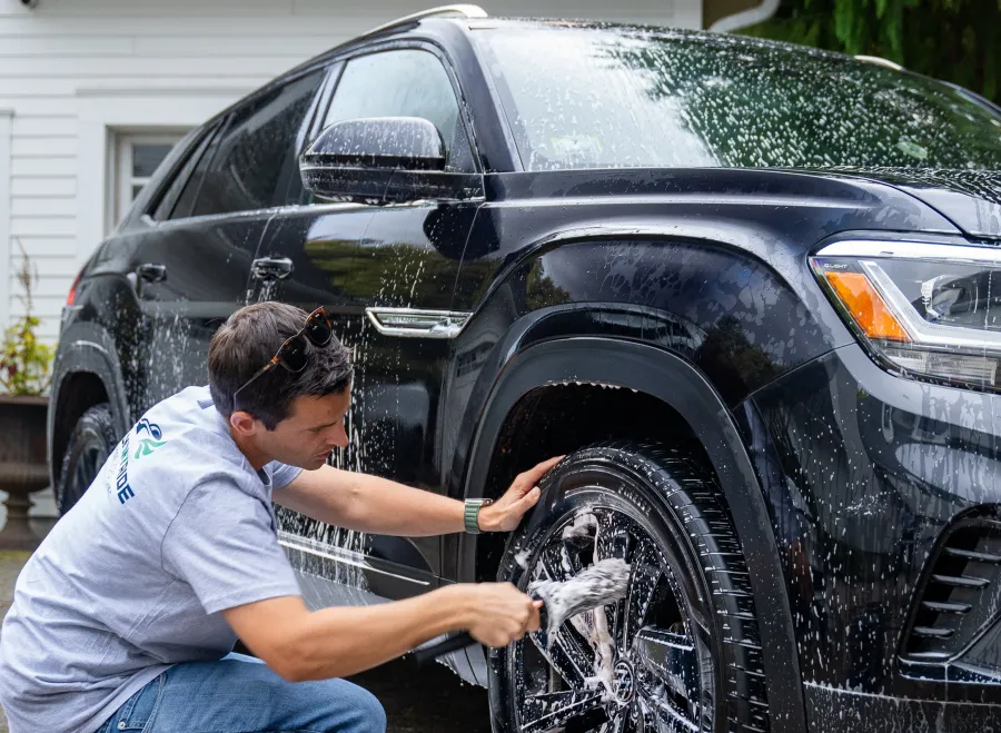 Man washing and scrubbing the front tire of a black SUV with soap suds in a residential driveway.