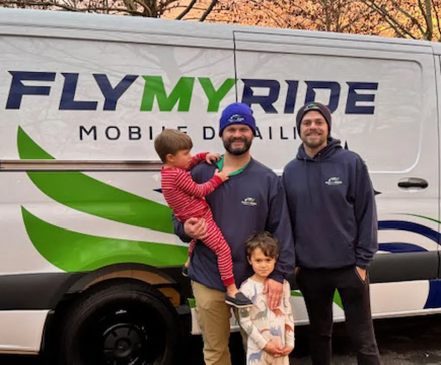 Two men and two children stand smiling in front of a Fly My Ride mobile detailing van with autumn trees in background.