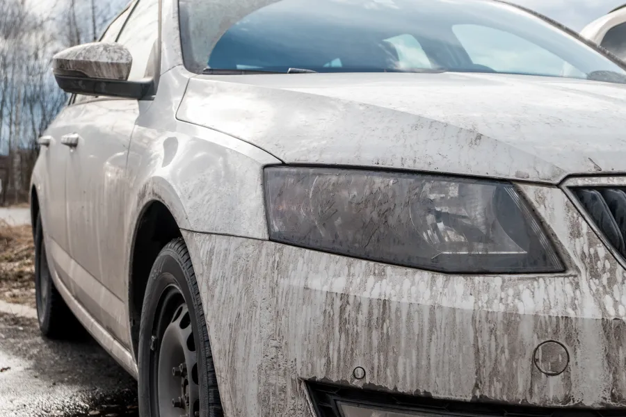 Close-up of a dirty white car with mud splattered on the front and side during daytime outdoors.