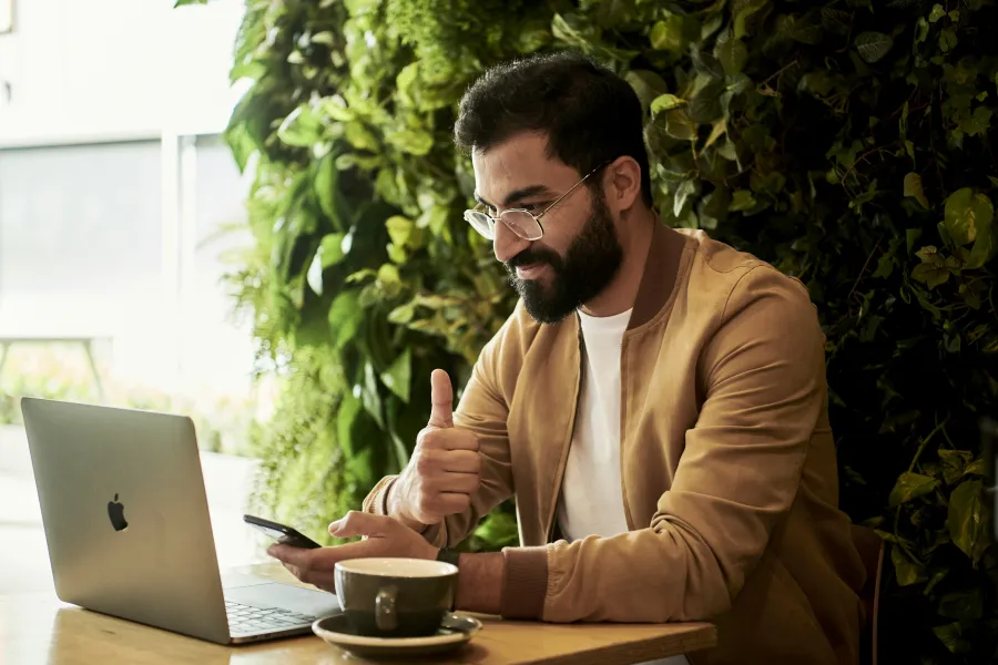 Man with glasses using smartphone and laptop, giving thumbs up while sitting at cafe table with coffee cup.