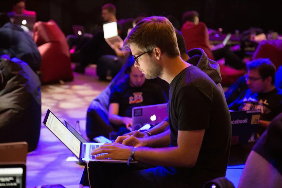 Young man focused on coding on laptop at a tech event with others working on laptops in a dimly lit room.