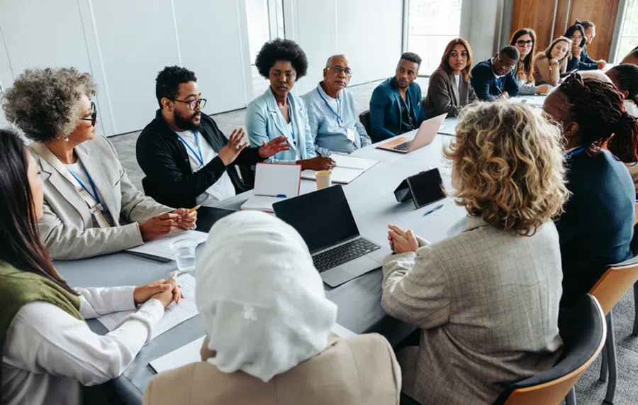 Diverse professionals engaged in a meeting around a conference table with laptops and notebooks.