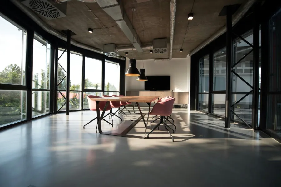 Modern conference room with large windows, concrete ceiling, wooden table, and pink chairs in natural light.