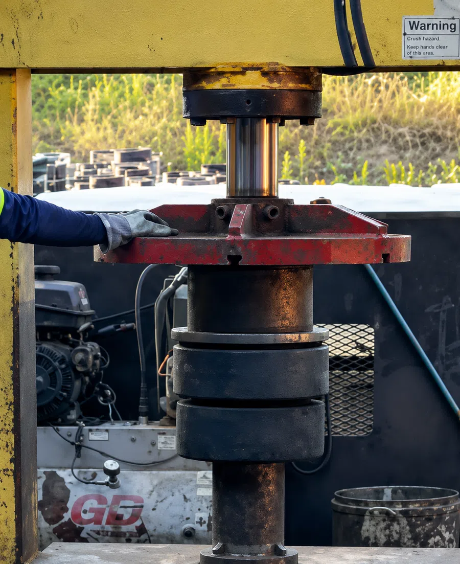 Worker's gloved hand operating hydraulic press machine in industrial setting with warning sign visible.
