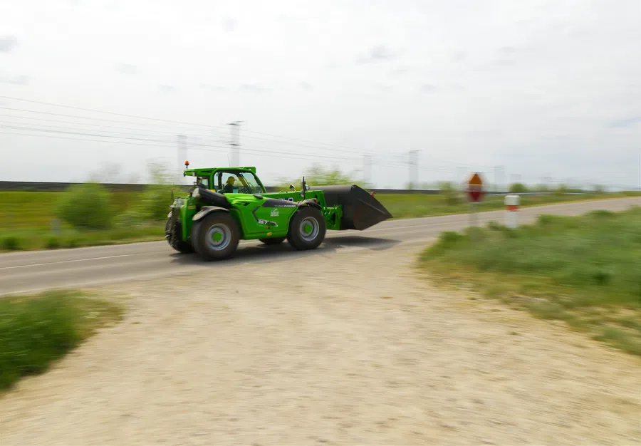 Green telehandler with bucket moving on rural road near grassy fields under cloudy sky